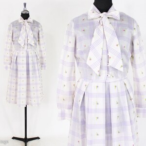 May include: A white dress with a purple and white checked pattern and small floral embroidery. The dress has a long sleeve, a bow tie at the neck, and a pleated skirt.