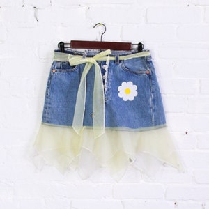 Puede incluir: Una falda vaquera con un volante de tul amarillo y un parche de margarita. La falda tiene una cinta amarilla atada a la cintura.