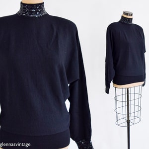 May include: Black mock neck sweater with sequined trim around the neck and cuffs. The sweater has a relaxed fit and long sleeves.