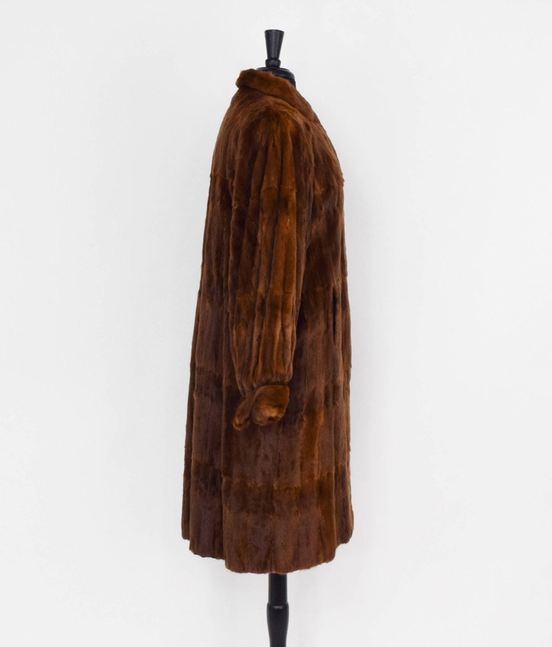 May include: A long brown fur coat with a collar and long sleeves. The coat is on a mannequin and is displayed against a white background.