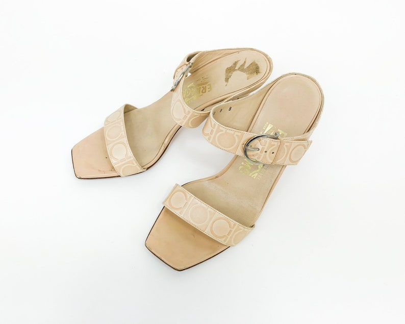 May include: A pair of beige leather heeled sandals with a buckle closure and a Salvatore Ferragamo logo pattern.