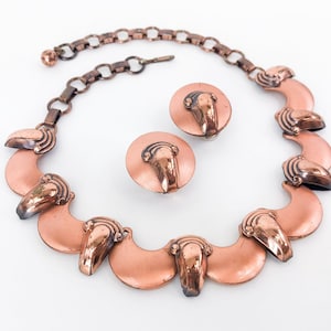 May include: A copper necklace and earrings set with a crescent moon design. The necklace has a chain and seven crescent moon pendants. The earrings are simple round discs with a crescent moon design.