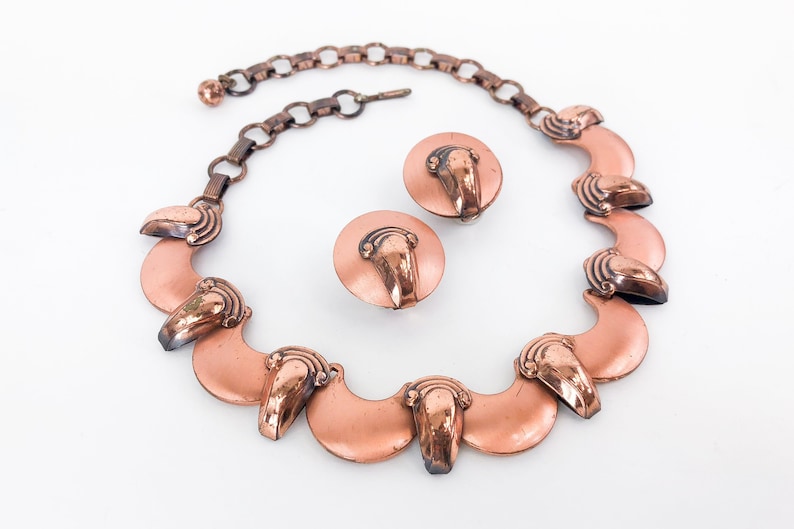 May include: A copper necklace and earrings set with a crescent moon design. The necklace has a chain and seven crescent moon pendants. The earrings are simple round discs with a crescent moon design.