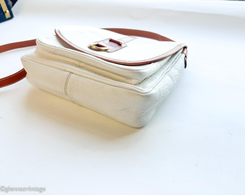 Puede incluir: Bolso bandolera de cuero blanco con correa y detalles en cuero marr&oacute;n. El bolso tiene un cierre de solapa con una hebilla dorada. Estilo vintage.
