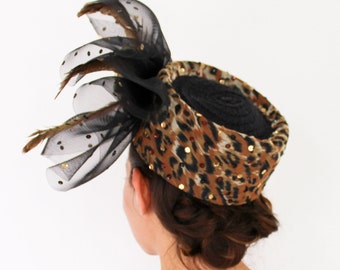 May include: A black and brown leopard print hat with a black net veil and feathers. The hat has a round shape and is decorated with gold sequins.