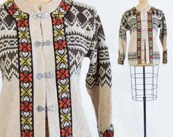 Puede incluir: Un cárdigan de punto beige con patrones geométricos marrones y blancos y detalles bordados de colores. El cárdigan tiene un cierre frontal con botones de metal plateado.