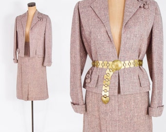 Puede incluir: Un traje de falda de tweed rosa malva con un cinturón de cadena dorada. La chaqueta tiene un cuello de muesca y mangas largas. La falda es de largo hasta la rodilla.