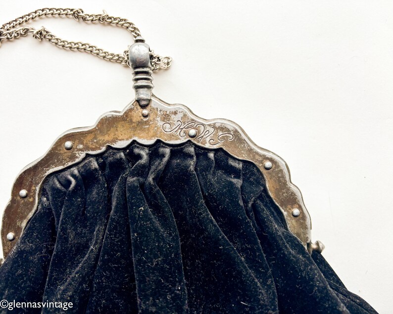 May include: A vintage black velvet purse with a silver metal frame and chain. The frame has an ornate design and the initials "M.W." are engraved on the front.