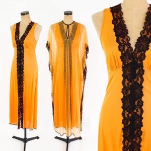 May include: Three vintage orange dresses with black lace trim are displayed on mannequins. The dresses include a sleeveless maxi dress and a sheer, flowing caftan. The black lace detailing is prominent along the neckline and down the front of the dresses.