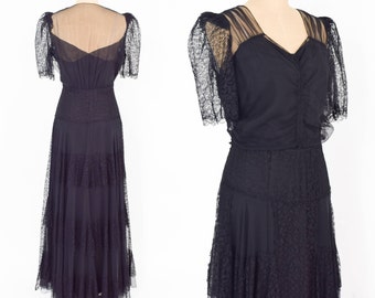 May include: A black floor-length gown with a sheer lace overlay and a tiered skirt. The dress has a V-neckline and short sleeves.