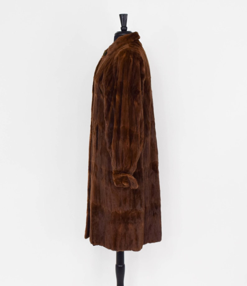 May include: A long brown fur coat with a collar and long sleeves. The coat is on a mannequin and is displayed against a white background.
