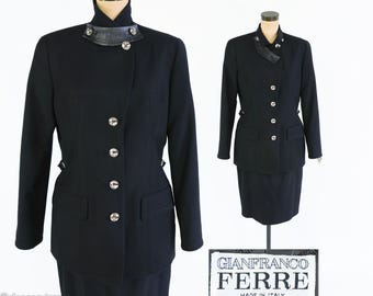 May include: A black two-piece suit with a jacket and skirt. The jacket features a high collar with leather detailing, silver buttons, and two front pockets. The skirt is a simple, straight cut. The suit is displayed on a mannequin.