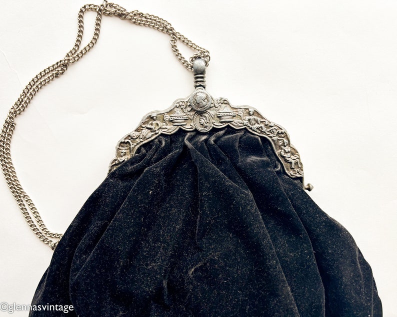 May include: A vintage black velvet evening bag with a silver chain and ornate metal clasp. The clasp features a detailed design with a central medallion.