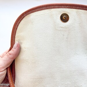Puede incluir: Primer plano del interior de un bolso vintage de cuero marr&oacute;n. El bolso presenta un interior de color crema claro con un cierre magn&eacute;tico dorado. Los bordes est&aacute;n ribeteados con cuero marr&oacute;n y costuras blancas.