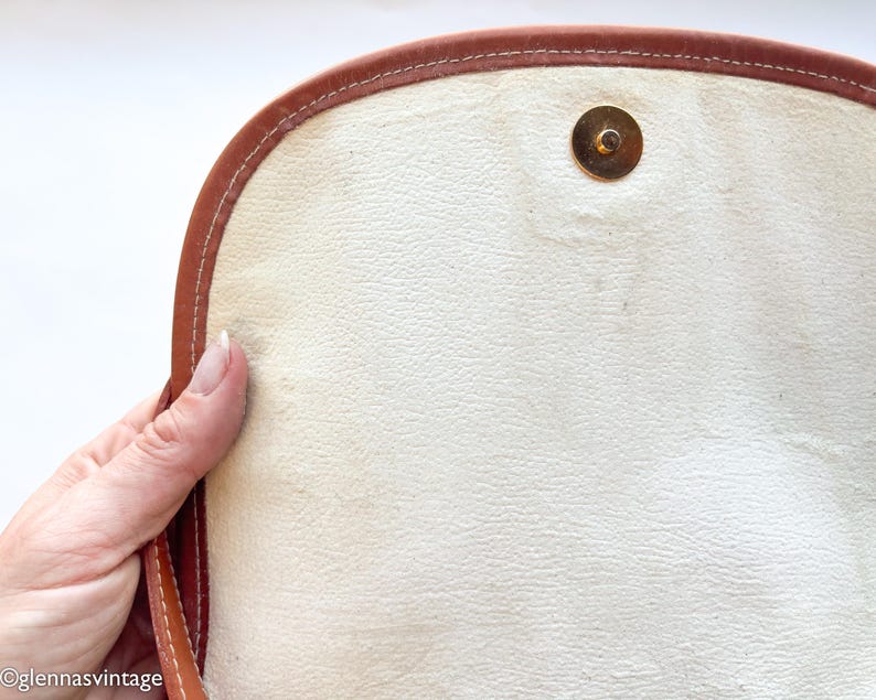 Puede incluir: Primer plano del interior de un bolso vintage de cuero marr&oacute;n. El bolso presenta un interior de color crema claro con un cierre magn&eacute;tico dorado. Los bordes est&aacute;n ribeteados con cuero marr&oacute;n y costuras blancas.