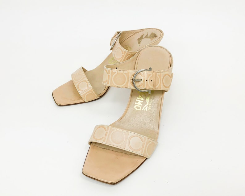 May include: A pair of beige leather heeled sandals with a buckle closure and the brand name "Salvatore Ferragamo" printed on the straps.