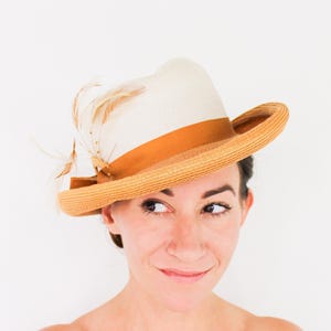 Può includere: Un cappello di paglia bianco e marrone con un nastro marrone e un accento di piuma. Il cappello è inclinato sulla testa di una persona con i capelli castani.
