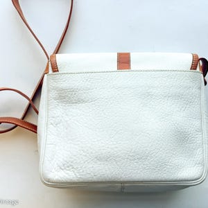 Puede incluir: Bolso bandolera de cuero blanco con detalles en cuero marr&oacute;n y una correa larga ajustable. El bolso tiene forma rectangular con cierre de solapa y hebilla dorada. El cuero texturizado del bolso a&ntilde;ade una est&eacute;tica vintage.