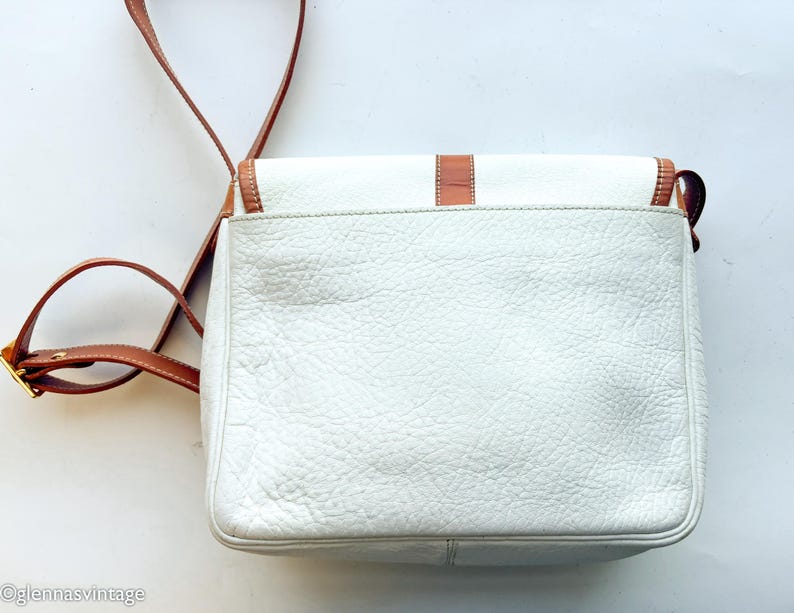 Puede incluir: Bolso bandolera de cuero blanco con detalles en cuero marr&oacute;n y una correa larga ajustable. El bolso tiene forma rectangular con cierre de solapa y hebilla dorada. El cuero texturizado del bolso a&ntilde;ade una est&eacute;tica vintage.