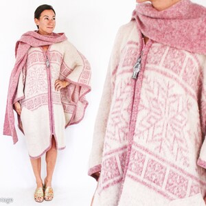 May include: A white and pink knitted poncho with a geometric pattern. The poncho has a zipper closure and a pink scarf is draped around the neck.