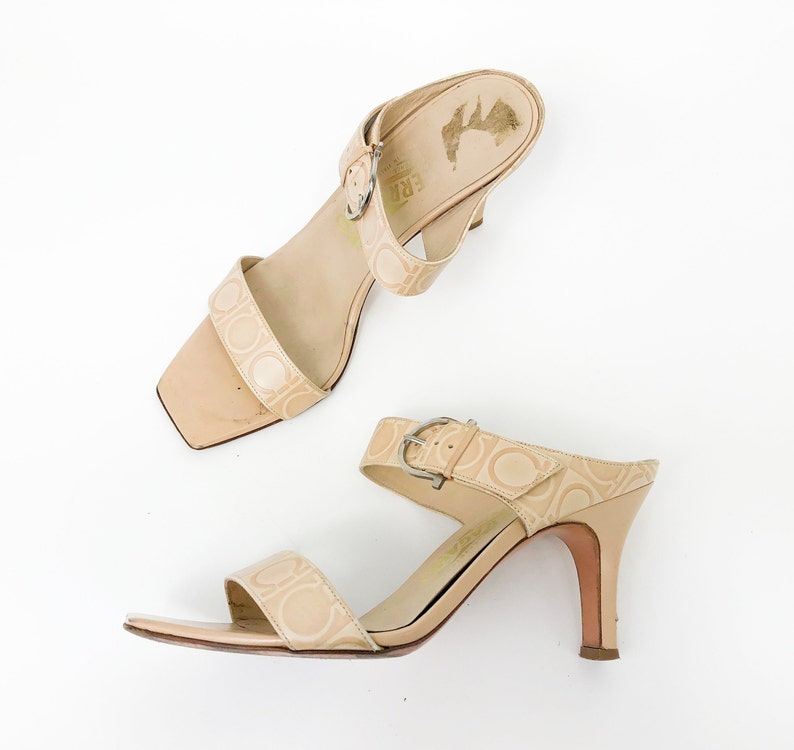 May include: A pair of beige heeled sandals with a buckle strap and a logo pattern. The sandals have a closed toe and an open heel.
