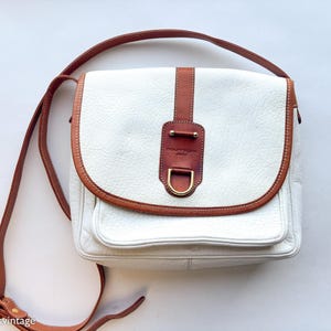 Puede incluir: Bolso bandolera de cuero blanco con ribete y correa de cuero marr&oacute;n. El bolso tiene un cierre de solapa con un adorno de cuero marr&oacute;n y una hebilla dorada. Tiene un bolsillo frontal y una correa larga ajustable.