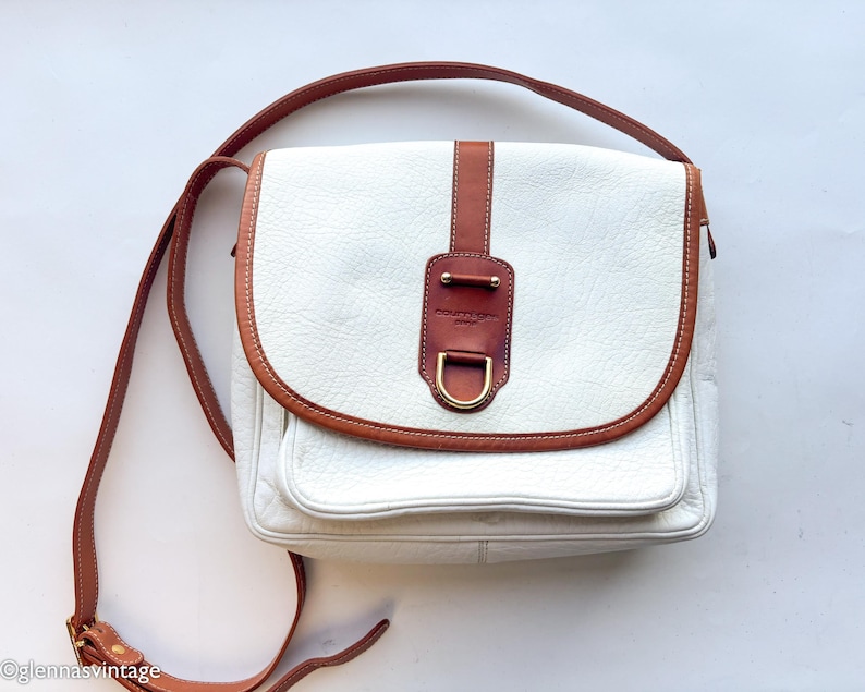 Puede incluir: Bolso bandolera de cuero blanco con ribete y correa de cuero marr&oacute;n. El bolso tiene un cierre de solapa con un adorno de cuero marr&oacute;n y una hebilla dorada. Tiene un bolsillo frontal y una correa larga ajustable.