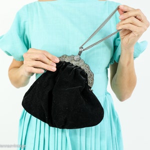 May include: A black velvet evening bag with a silver metal clasp and chain strap. The bag is closed and has a gathered, pleated look.