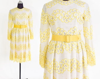 Puede incluir: Un vestido blanco con detalles de encaje amarillo y un cinturón amarillo. El vestido tiene mangas largas y un corpiño ajustado. La falda es amplia y fluye desde la cintura.