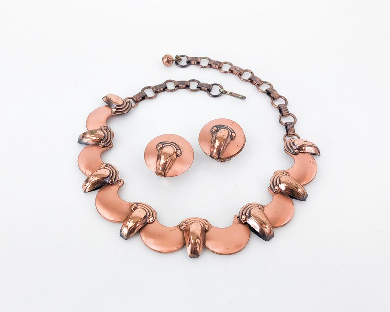 May include: A copper necklace with a chain and matching earrings. The necklace features a series of crescent-shaped pendants with a decorative design.