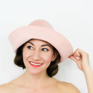 May include: A woman wearing a light pink wide-brimmed hat. The hat is made of felt and has a rounded crown.