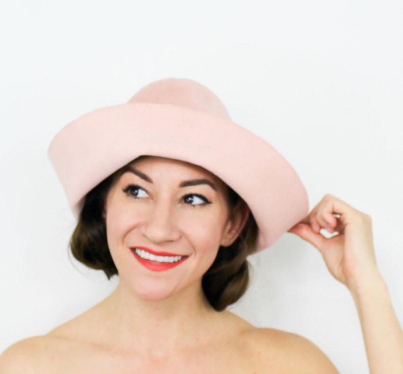 May include: A woman wearing a light pink wide-brimmed hat. The hat is made of felt and has a rounded crown.