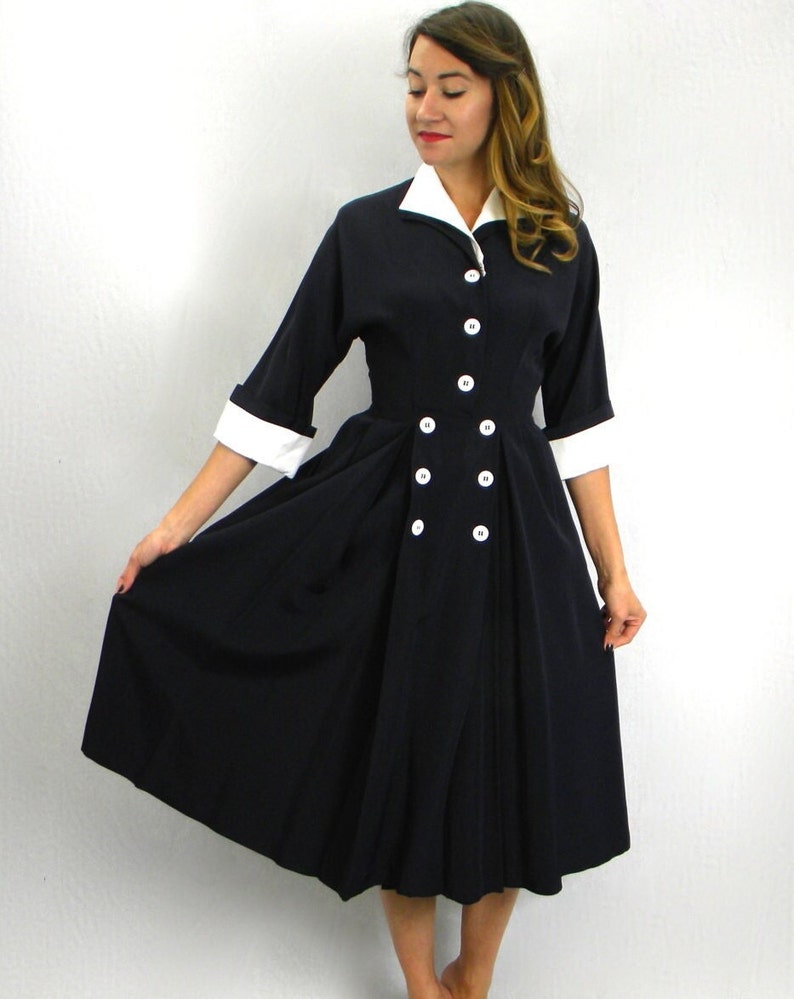 Pode incluir: Um vestido azul marinho com colarinho e punhos brancos. O vestido tem um fecho de bot&otilde;es na frente e uma cintura ajustada. A saia &eacute; ampla e chega aos joelhos.