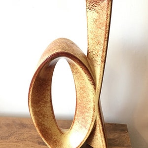 May include: A tan and brown ceramic vase with an abstract design. The vase features a large loop and a tall, slender section. The vase is on a wooden surface.