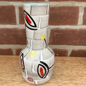 May include: A ceramic vase with a light blue interior and a white exterior featuring a grey grid pattern. The surface is decorated with black outlined leaf shapes containing red centres, along with scattered yellow and pink circular accents.