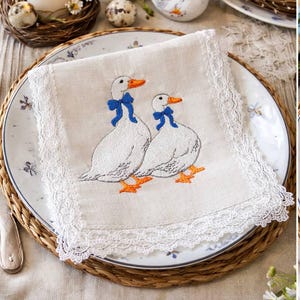 Può includere: Un tovagliolo di lino bianco con oche ricamate, ognuna con un fiocco blu e zampe arancioni, è posto su un piatto decorativo. Il tovagliolo è bordato con pizzo bianco. La scena comprende una candela, uova e una statuina di oca in ceramica.