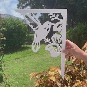 Op de afbeelding: Witte metalen tuinpaal met een kolibrie en een bloem. De kolibrie is in de vlucht met uitgespreide vleugels. De bloem is een hibiscus met vijf bloemblaadjes.
