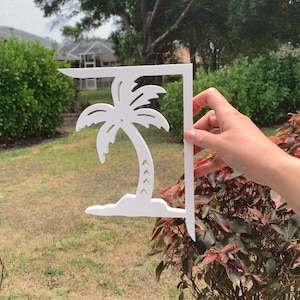 May include: White wooden palm tree cutout with a simple design. The cutout is attached to a white wooden bracket. The bracket is held by a hand.
