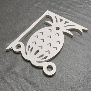 Op de afbeelding: Wit houten ananas-plankensteun met een decoratief ontwerp. De steun heeft een gebogen onderkant en een rechte bovenkant met een gat voor montage.