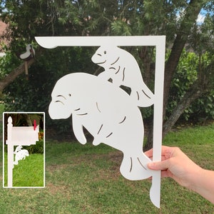 Op de afbeelding: Witte houten brievenbusbeugel met een uitgesneden ontwerp van twee lamantijnen. De grotere lamantijn bevindt zich onderaan en de kleinere lamantijn bevindt zich bovenop de grotere. De beugel wordt vastgehouden door een hand.