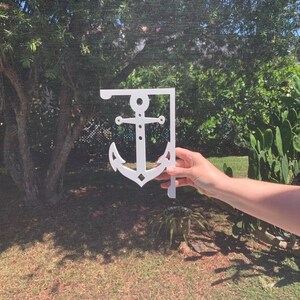 May include: White metal anchor-shaped shelf bracket. The bracket is attached to a wall and is being held by a hand.
