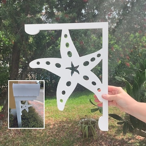 Può includere: Un palo di cassetta postale bianco con un design a stella marina bianca. Il palo è attaccato a una cassetta postale bianca. La cassetta postale è sullo sfondo dell'immagine. Il design a stella marina è in primo piano dell'immagine.