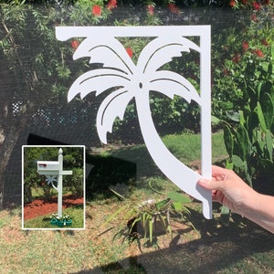 May include: A white, decorative mailbox bracket featuring a cut-out palm tree design. The bracket is held up in front of a green background. The image also shows a white mailbox with a palm tree design.
