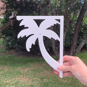 May include: White wooden palm tree shelf bracket. The bracket is shaped like a palm tree with a curved trunk and fronds. It is a decorative shelf bracket that can be used to add a tropical touch to any room.