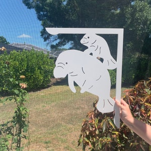 May include: A white L-shaped decorative bracket featuring a cutout design of an adult manatee with a smaller calf manatee positioned above it. The bracket has a flat top edge and a vertical side edge, with the manatee silhouettes integrated into the frame.