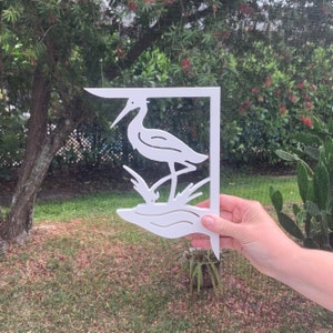 Puede incluir: Soporte de pared de metal blanco con un diseño recortado de una garza de pie en el agua. El soporte está diseñado para ser utilizado como elemento decorativo para un jardín o un hogar.