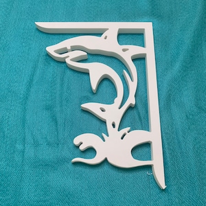 Puede incluir: Soporte de estante decorativo blanco con diseño de delfín. El soporte presenta un diseño recortado de un delfín saltando fuera del agua. El soporte está hecho de material blanco sólido y está diseñado para montaje en pared.