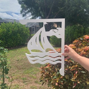 Op de afbeelding: Witte metalen wandbeugel in de vorm van een zeilboot met een uitgesneden ontwerp. De zeilboot vaart op golven en heeft een zeil dat in de wind waait.