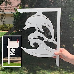 May include: White wooden mailbox bracket with a dolphin design. The bracket is shaped like a wave with two dolphins jumping out of the water. The bracket is mounted on a white wooden post.