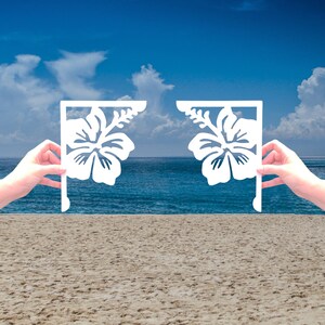 Könnte beinhalten: Zwei weiße Metall-Eckkonsolen mit einem Hibiskus-Blütenmuster. Die Konsolen werden vor einer Strandkulisse mit blauem Wasser und Sand gehalten.